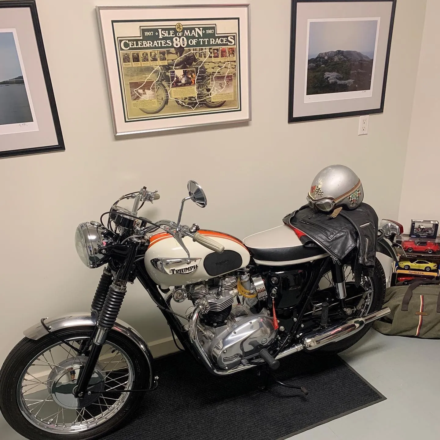 Pristine vintage Triumph motorcycle with white and orange fuel tank in a collector's garage with Isle of Man TT race memorabilia on the wall