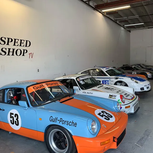 Row of vintage Porsche race cars including a classic Gulf-livery 911 parked inside a professional speed shop